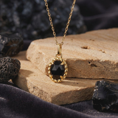 Close-up of the solid gold necklace and oval black onyx pendant resting on stacked tan stone blocks.