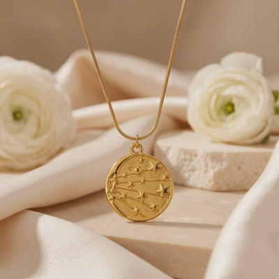 Close-up of a gold-tone celestial medallion necklace displayed on cream fabric beside white flowers.