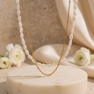 Layered gold-finish necklace displayed above a round stone pedestal, with white flowers in the background.