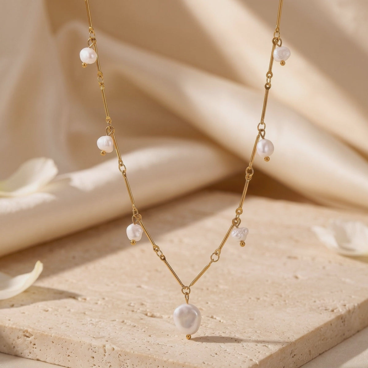 Gold-tone necklace with irregular white pearls displayed on a beige stone slab with draped fabric behind it.