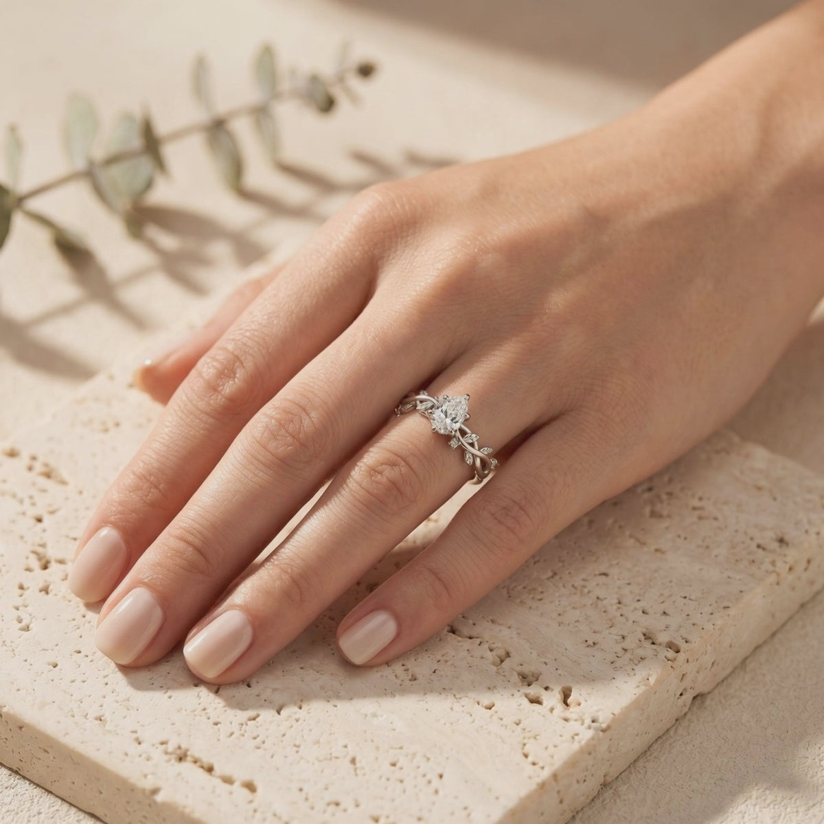Solid gold leaf-vine band ring with pear-cut moissanite worn on a hand over beige stone tile.
