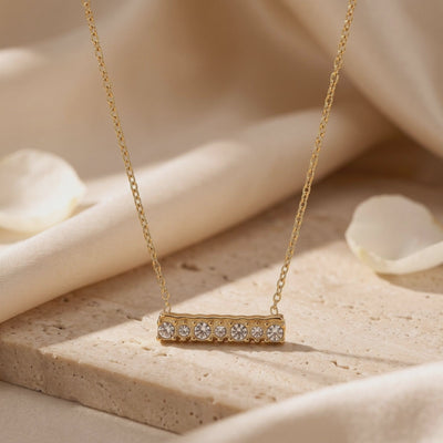Close-up of the gold-toned bar necklace with seven round clear stones on a textured beige block.