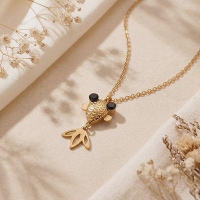Gold-tone goldfish pendant necklace with black eyes arranged on cream fabric beside dried flowers.