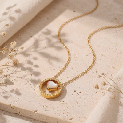 Gold-toned necklace with a mother-of-pearl heart inside a circular pendant, arranged on a textured stone surface.