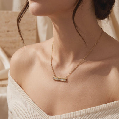 Model wearing a gold-toned horizontal bar necklace with seven clear stones on a cream ribbed top.