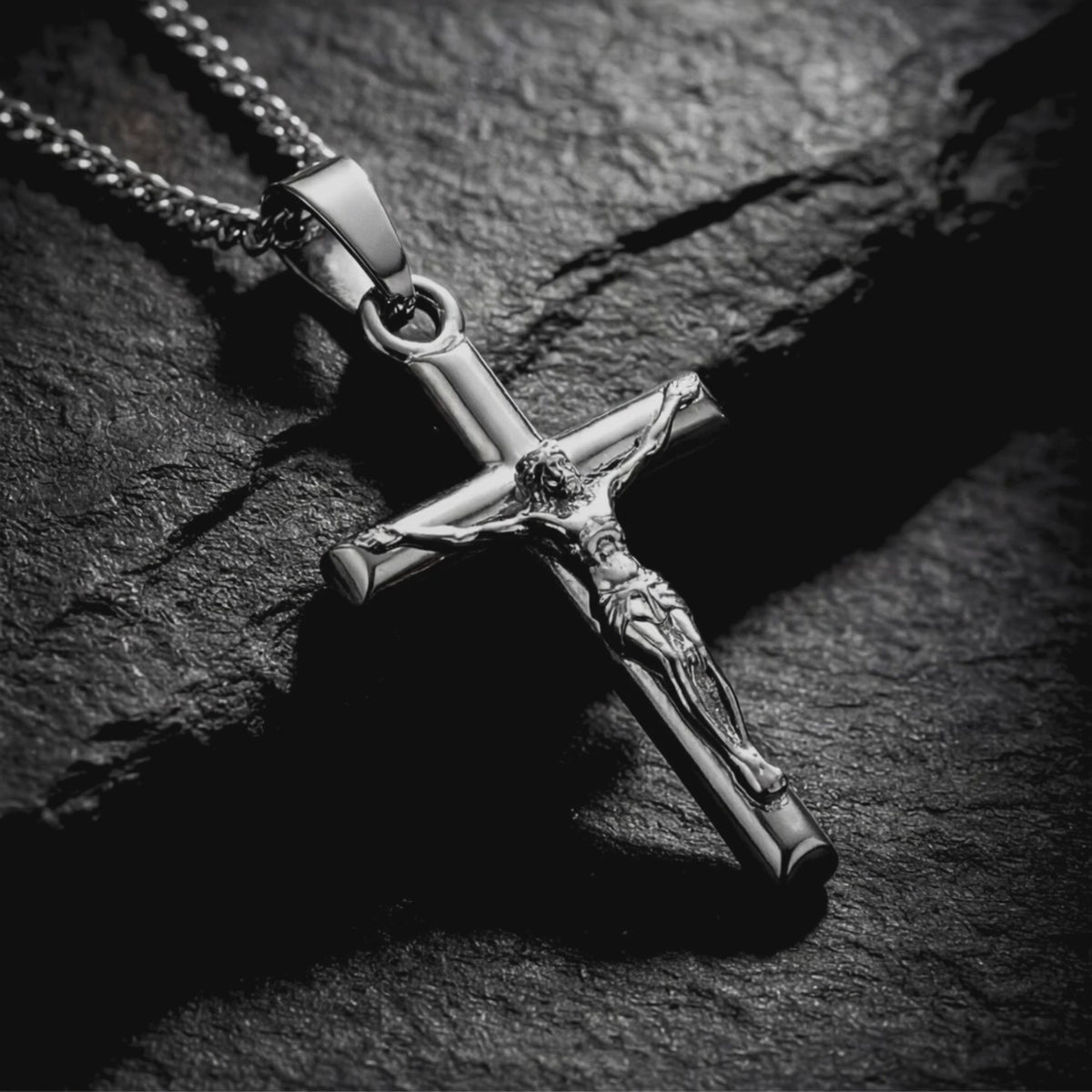 Close-up of a silver crucifix pendant and chain resting on a dark textured stone surface.