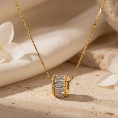 Close-up of the gold-tone cylindrical pendant with clear baguette-cut stones on its chain over a stone surface.