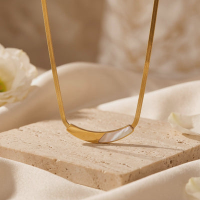Gold snake chain necklace with a curved mother-of-pearl bar, displayed on a textured stone block.